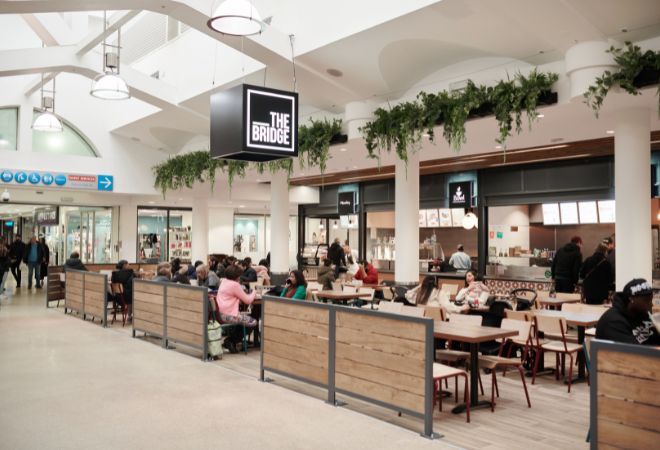 nterior of a spacious food court with natural lighting. Multiple wooden tables with attached seating are occupied by various individuals and groups. A large, hanging sign reads “THE BRIDGE” above a serving area with menu boards. Decorative greenery hangs from the ceiling, adding a touch of nature to the setting.
