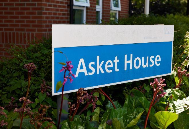 Blue and white sign that reads 'Asket House' surrounded by greenery and plants with a brick building behind.