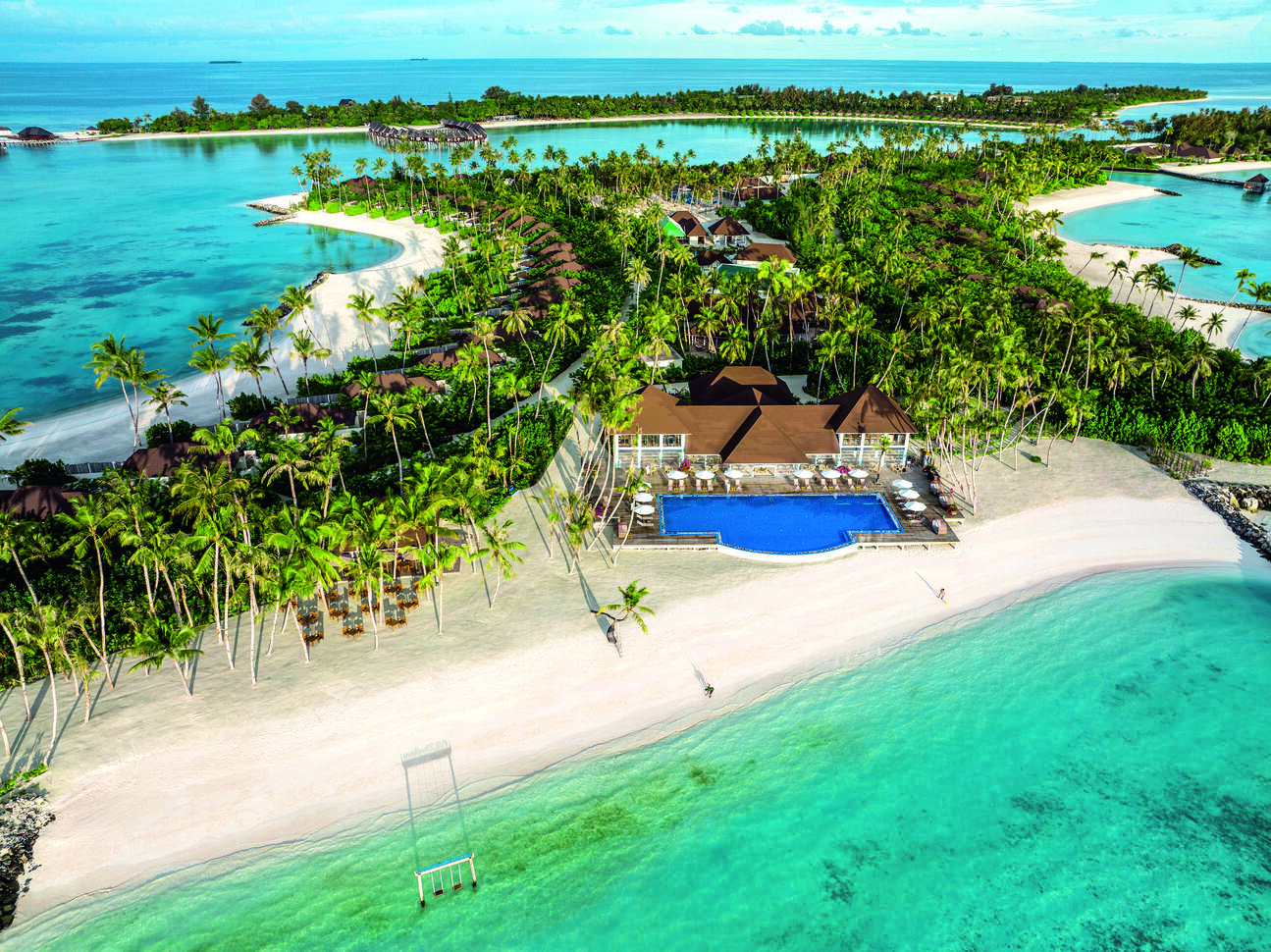Aerial view of the resort right on the golden sandy beach with the clear blue sea in front. Surrounded by a forest of palm trees and sea all around the small island.