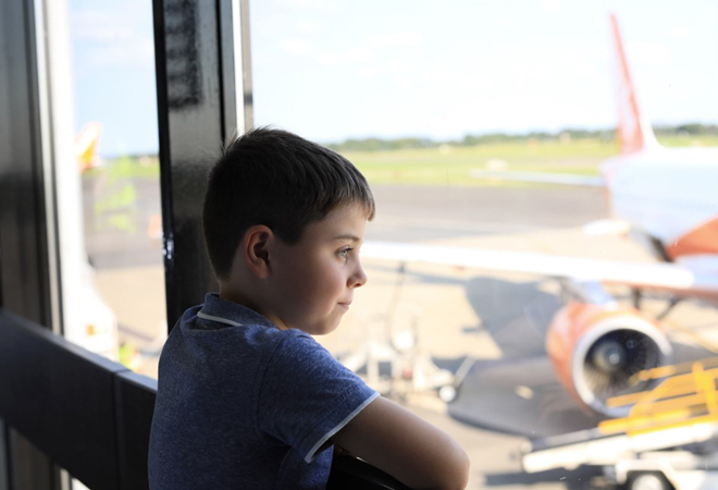 "Photograph of a white male child in blue t shirt in the departures area, looking out a window towards aircraft outside"