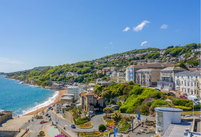 "Sunny blue skies and an aerial shot of the coast with houses, shops, a winding road and mountains in the background"