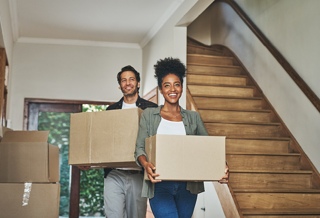 "a couple are carrying boxes inside a house, with stairs behind them. some more boxes are on the floor"