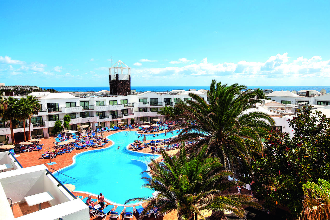 Aerial view through the palm trees of the resort with the large pool with people in and around on sun loungers, and the white hotel buildings behind with the sea visible in the distance on a slightly cloudy but sunny day.