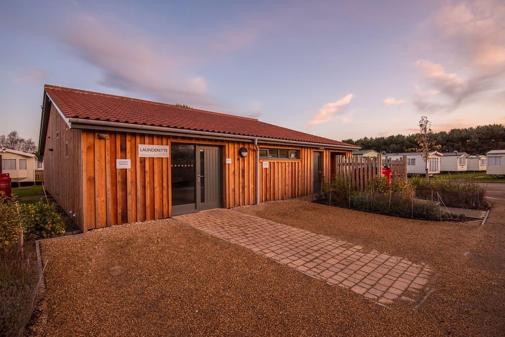 Wooden laundrette building with red roof at sunset, surrounded by greenery and mobile homes. The sky is partly cloudy, casting a warm glow over the scene.