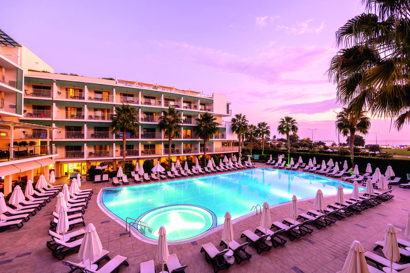View in the dusk of the pool area surrounded by many sun loungers and umbrellas with clusters of hotel rooms behind and palm trees dotted around.