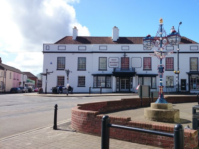 "sally mitchell's gallery is across the road, a white building. a signpost is in the foreground, with two intricate lamps on either side."