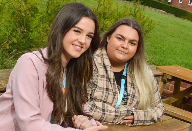 "Photo of 2 women sat at a picnic bench smiling for a photo. The person on the left has long brown hair and a pink jacket on. The person on the right has long blonde hair and a brown check shirt."