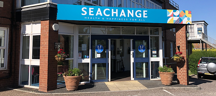 An entranceway with a large blue banner stating 'Seachange health & happiness for all', hanging plant pots surround the blue doorway, as well as large white windows around the doors