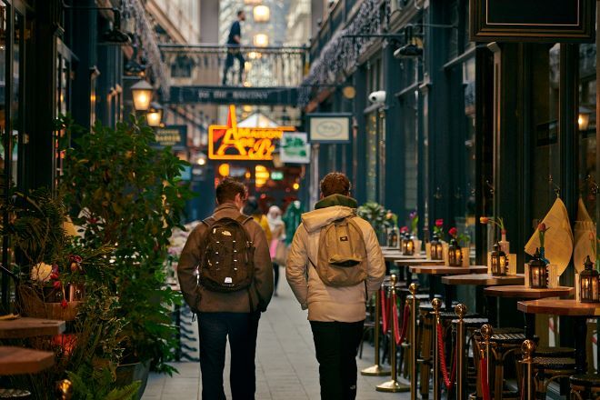 "Two people walking away through Cardiff Arcade, restaurant seating to either side"