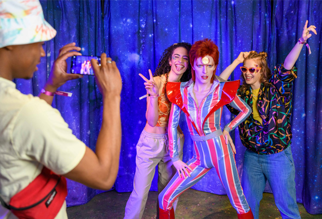 "A person taking a photograph of two people stood posing with a wax figure of David Bowie dressed in a blue and red PVC jumpsuit"