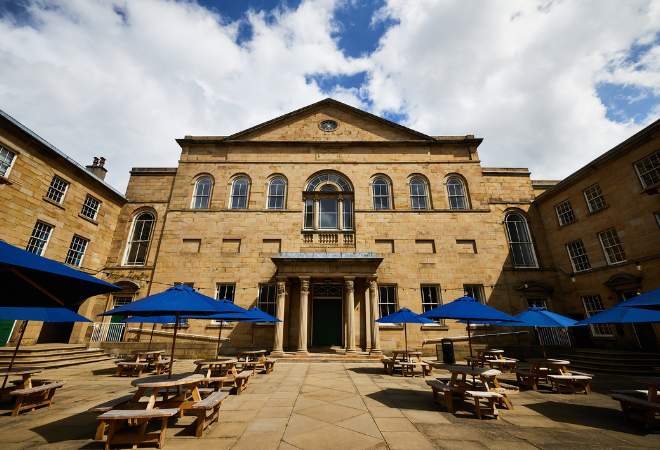 The exterior of the theatre is a light coloured brick with a large entrance with 4 pillars around the door, there are some small steps leading up to the door and either side of the entrance there is wheelchair ramps. There is one large window above the door and then smaller windows to the sides. The building goes around each side in a U shape with a patio out the front with many picnic benches and blue umbrellas.
