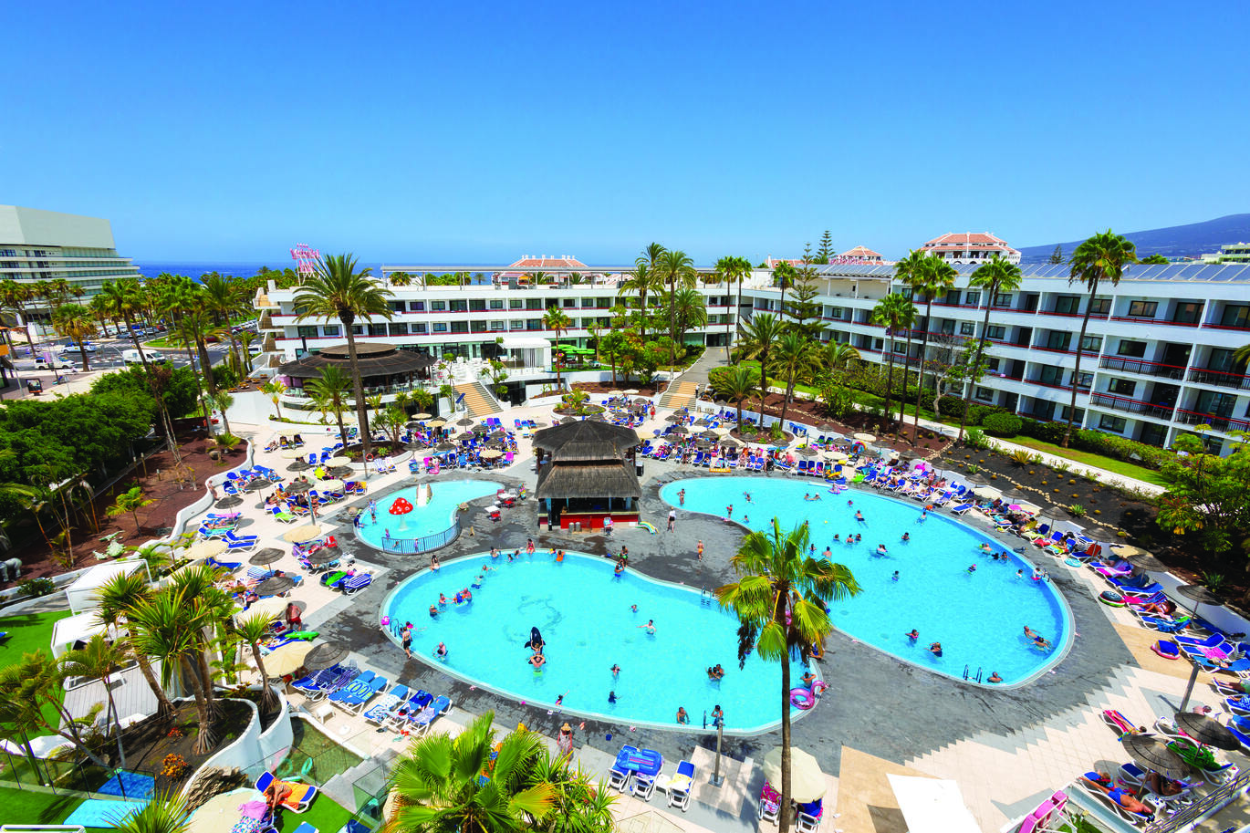 Aerial view of the hotel complex with 3 pools in the middle and the hotel building surrounding. With many people in and around the pool on sun loungers on a clear sunny day.