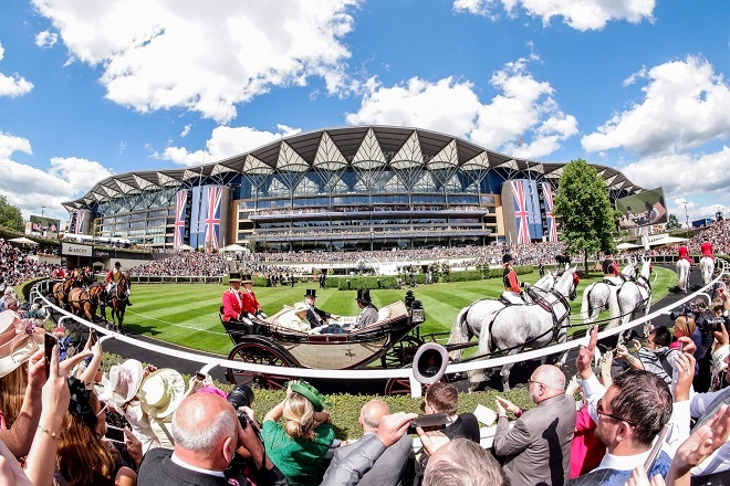 "Photo at Royal Ascot. There are crowds at the bottom of the pitching looking out onto the course. On the course there are many horse drawn carries carrying people. In the background is one of the stands for ascot with many people in them. There are British flags displayed to the sides of the stands"