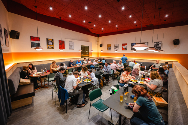 An indoor dining area with multiple groups of people seated at various tables. The room has a red ceiling, white walls adorned with framed pictures, and a combination of booth seating along the perimeter and freestanding tables in the centre. Overhead lighting fixtures illuminate the space