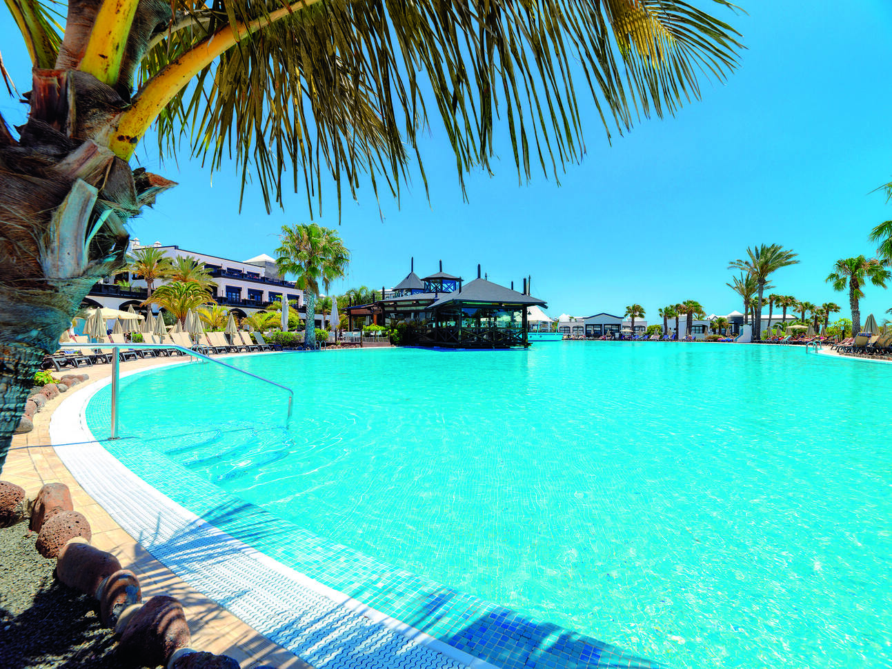 A view of the lush blue pool with the hotel building surrounding. There are some beige sun loungers and umbrellas either side of the pool and a number of palm trees around. It is a lovely sunny clear day.
