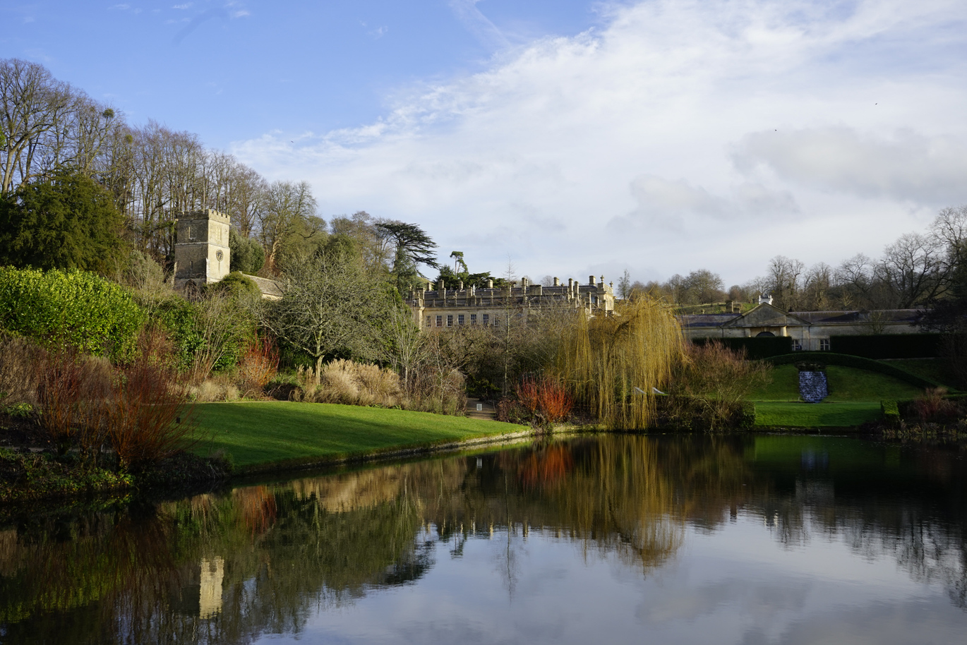 A large body of water surrounded by a lush green lawns with a church and large building peeking out from behind the trees.