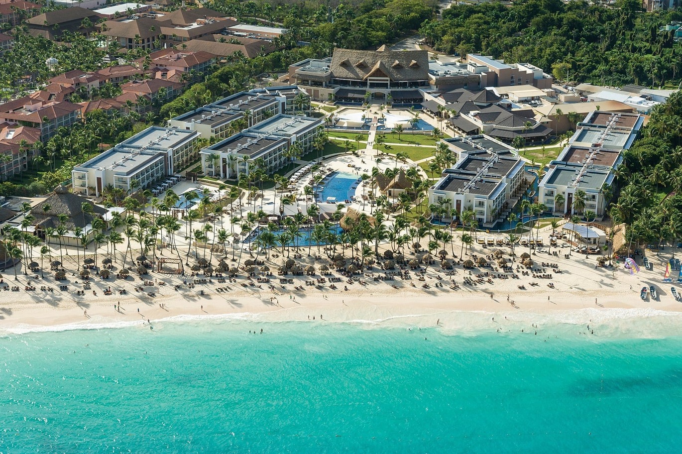 Aerial view above the beautiful greeny blue sea and lots of people dotted around the white beach. Palm trees are directly behind with the hotel complex then behind which is surrounded by forests of trees and a few different building.