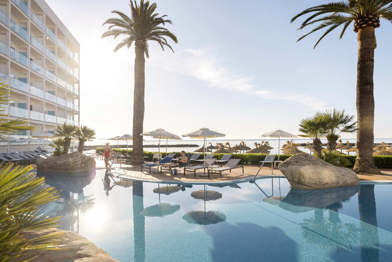 At the front of the picture is the lovely blue pool with a woman in a red swim suit getting in. Around the pool are some large rocks and some sun loungers with umbrellas over them where a man is laying reading a book. There are 2 large palm trees just by the pool. To the left you can see the hotel building with a cluster of hotel rooms and balconies. Directly behind the pool is the beach where you can see a number of straw like umbrellas and the sea behind them.