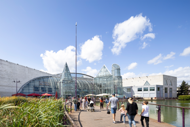 "Exterior view of Bluewater shopping centre with glass-domed entrance, lakeside boardwalk and people walking."