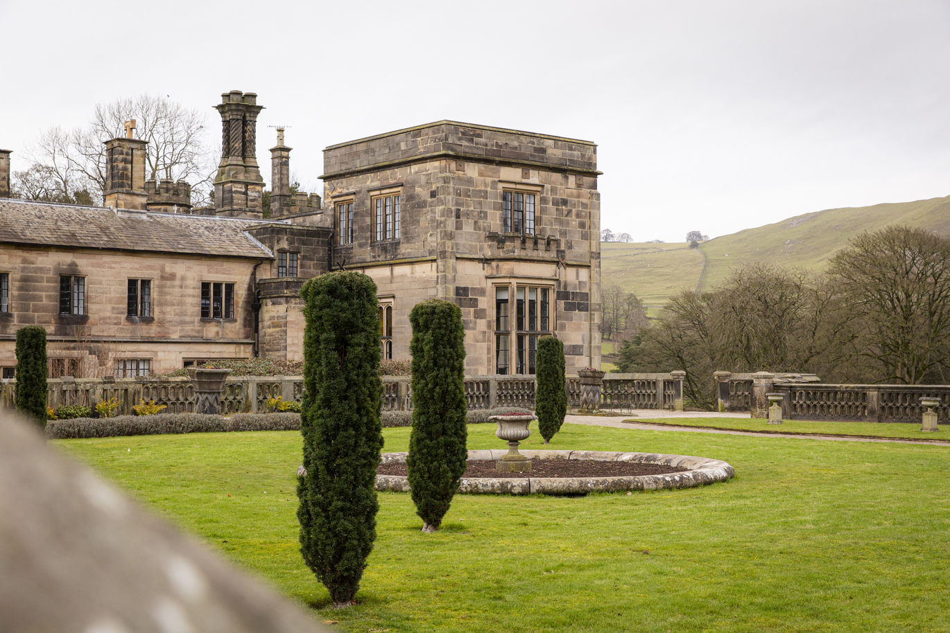 A large stone 16th century building with trees and hills behind, with a lush green lawn out the front with a stone fountain in the middle. There are a number of tall, thin, neat hedges around the lawn.