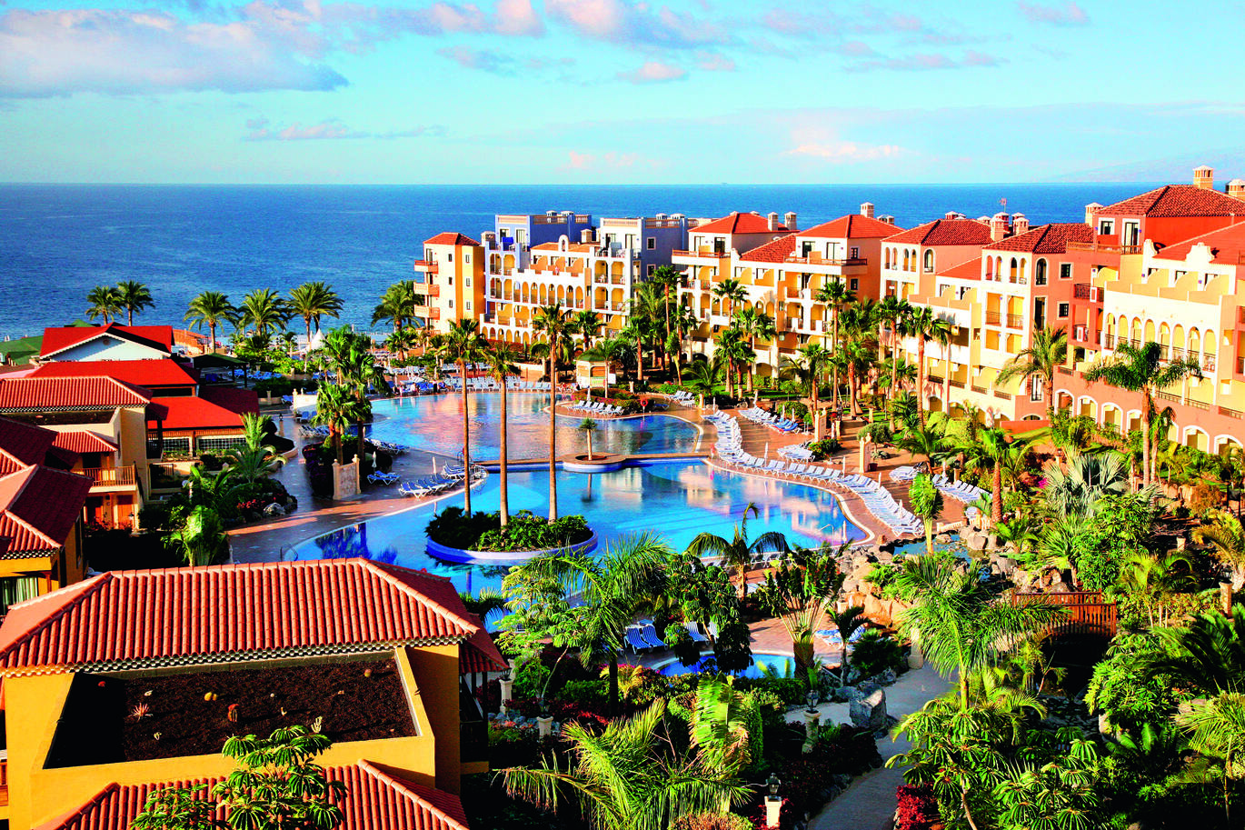 Aerial view of the hotel complex with the pool in the middle surrounded by the hotel buildings and many palm trees, with the sea visible behind.