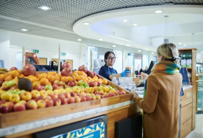 An indoor scene of a grocery store with a focus on the fruit section. There is a display of various apples in the foreground, with other produce items slightly out of focus in the background. A person with their back to the camera is standing in front of the apple display, seemingly making a selection or examining the fruit. Another individual is partially visible behind a checkout counter in the midground, facing away from the camera and obscured by pixelation for privacy.