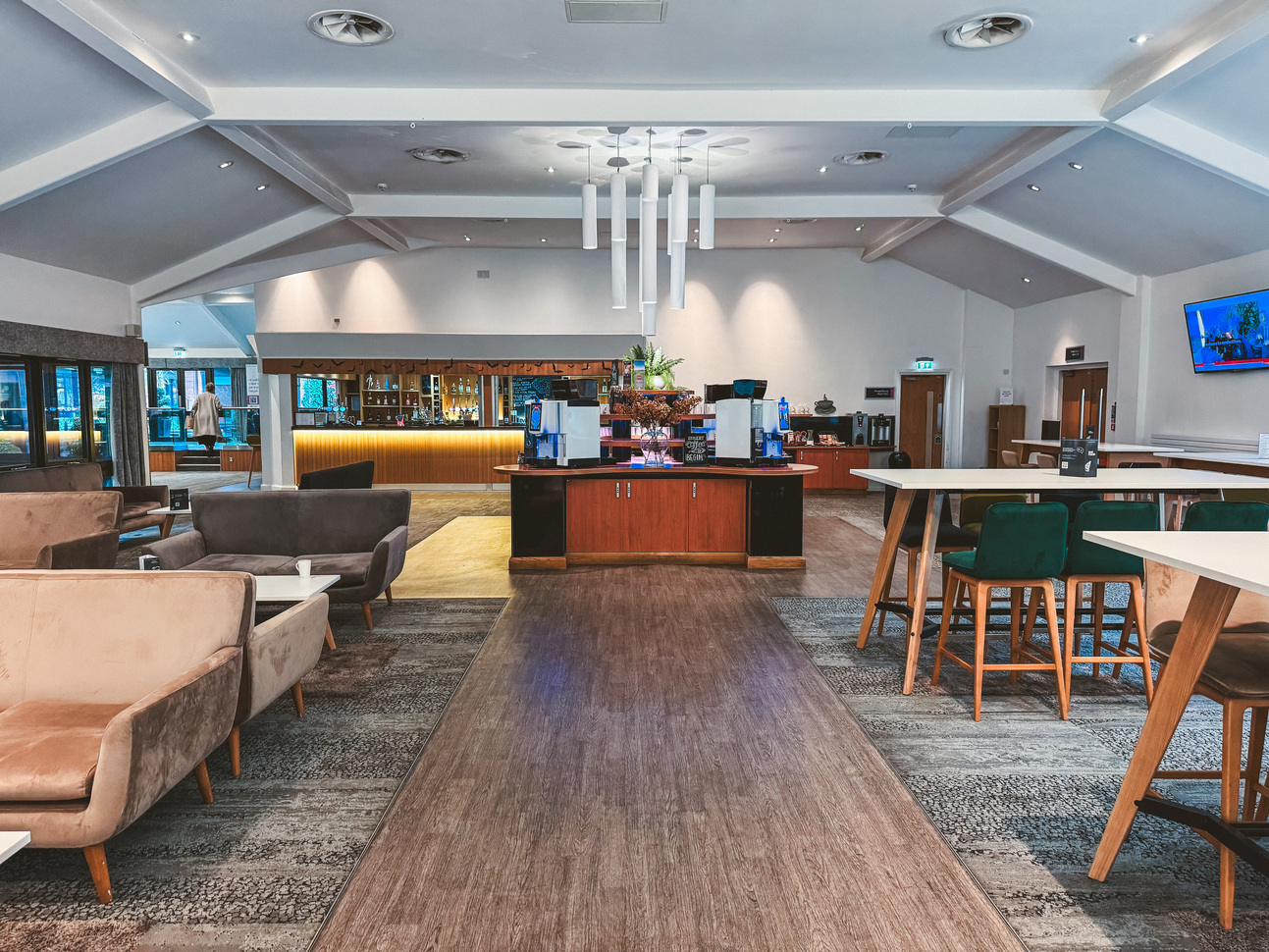 "Lounge area with wooden floored walkway, low grey and brown sofas with white coffee tables on the left of the walkway, high up green and brown stools with white tables on the right, a bar/coffee station at the back of the photo"