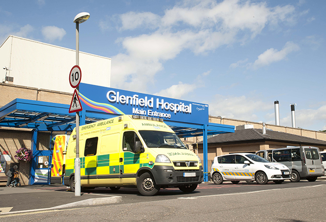 "External of Glenfield hospital main entrance, an ambulance is parked in the foreground with its rear doors open."