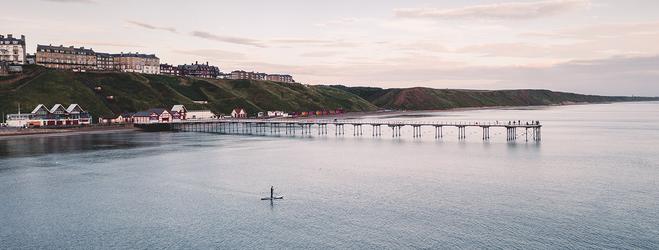 Coastal view of a seaside town with a long pier extending into calm water, cliffs and buildings along the shoreline, and a lone paddleboarder on the bay.