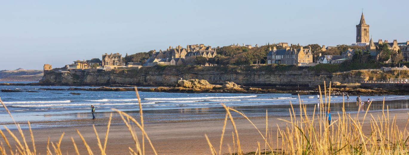 "Beach with small rolling waves. Long grass in foreground. St Andrew's town in background set against clear blue skies."