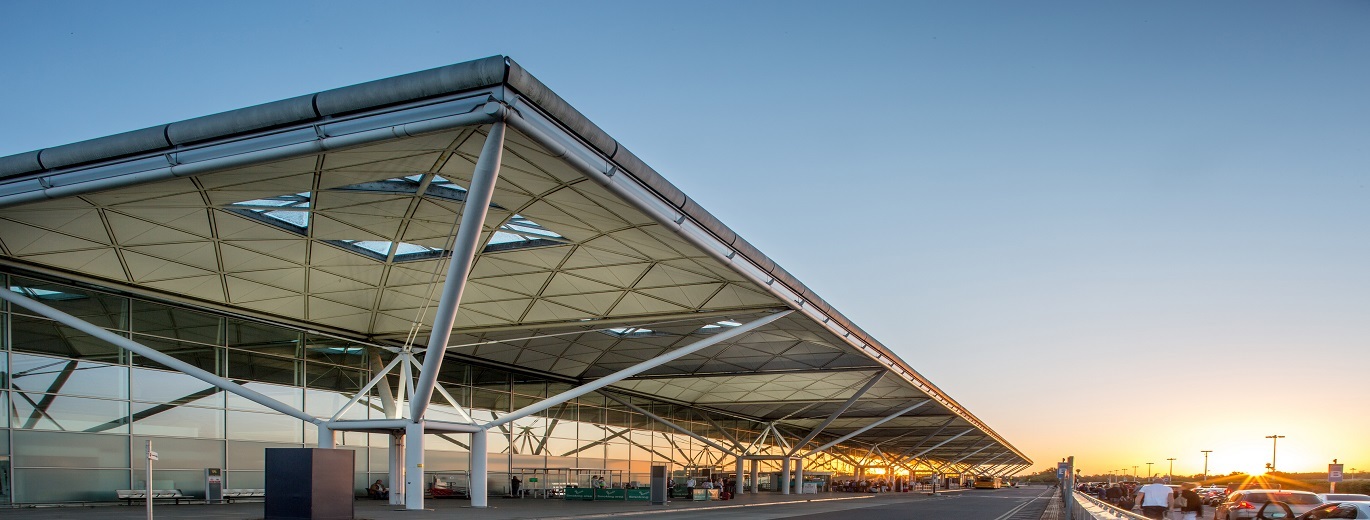 "External photo of London Stansted Airport terminal building. There is parking to the right of the photo, a road in the middle and the building to the left. There is a large roof covering the path the full length of the building in the photo."