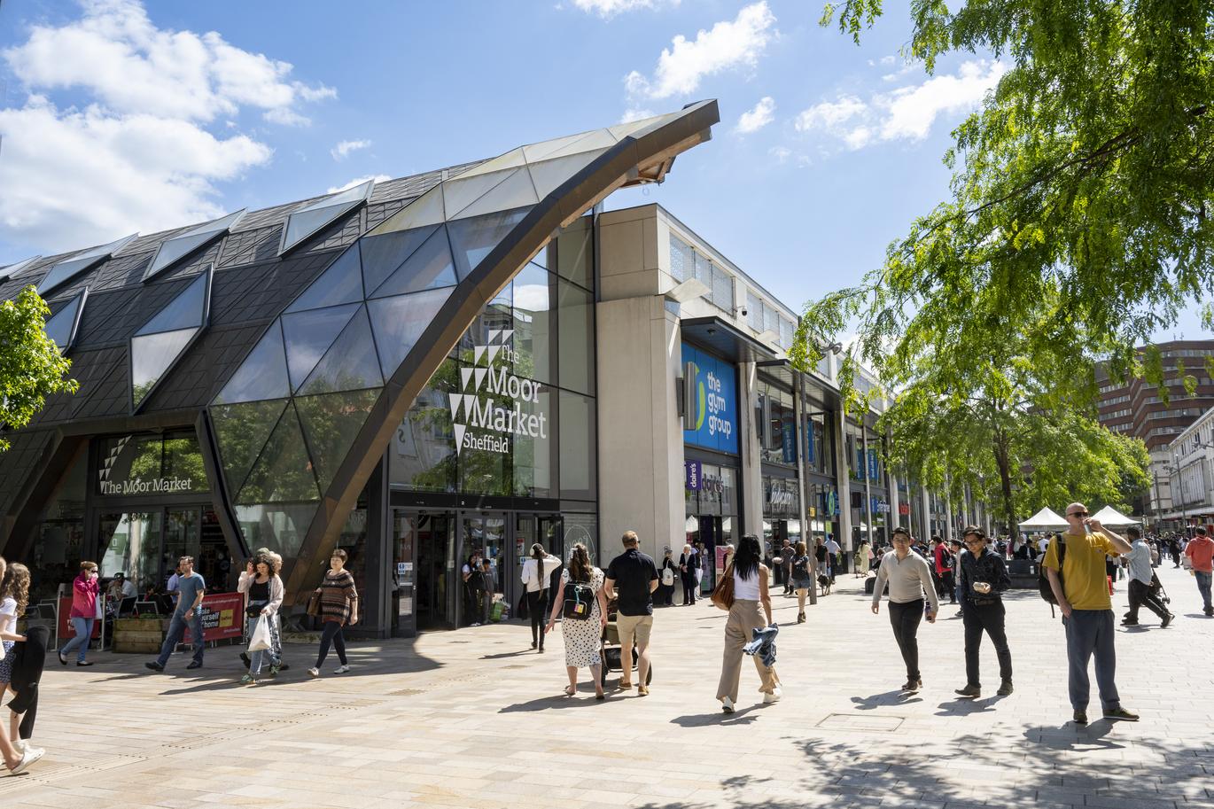 "Outside of The Moor Market, Sheffield" on a sunny Spring day with people walking in the sunshine including a couple pushing a pram, with glimpses of a tree on both the far right hand side and far left hand side of the photograph"