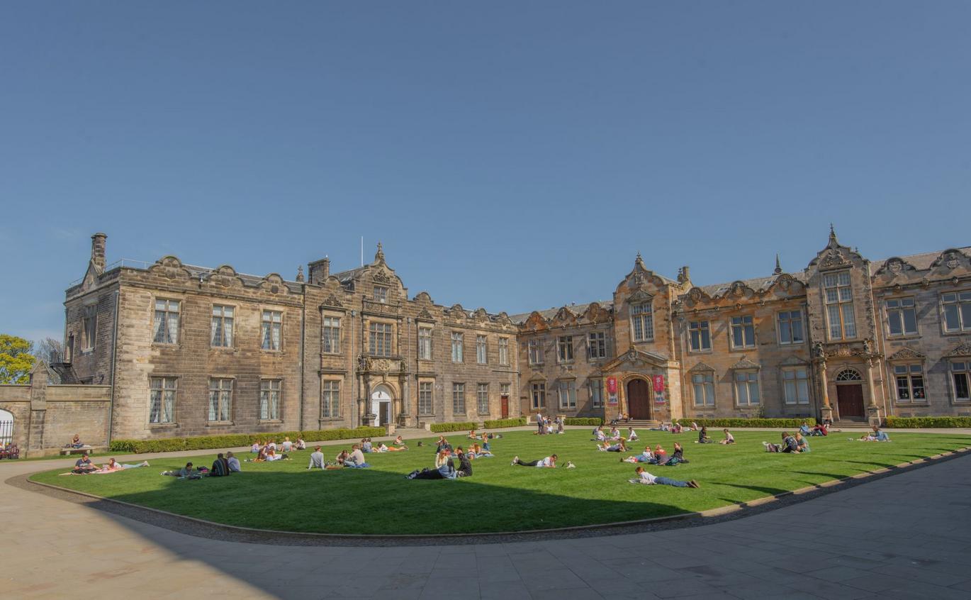 “Sunny day at a historic university quadrangle with a large green lawn surrounded by ornate stone buildings. People are sitting and lying on the grass relaxing, studying, and socialising under a clear blue sky.”