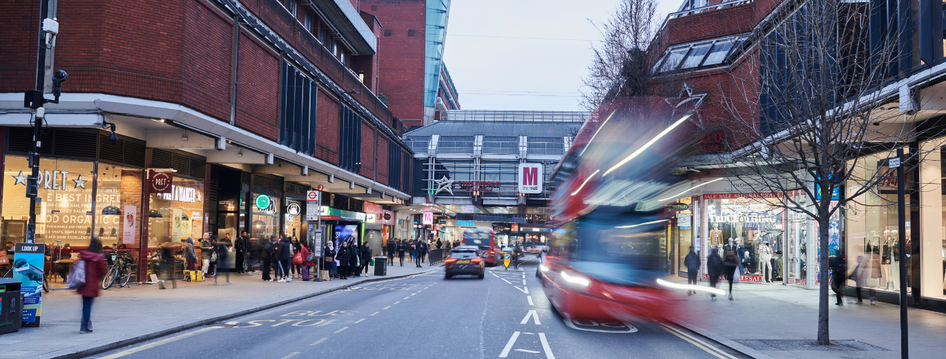A panoramic view of a busy urban street scene at dusk with blurred motion of a red bus moving, indicating the bustling city life. The street is lined with various shops and pedestrians, and cars are visible in the distance. 