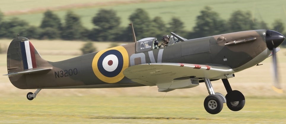 "Photo of a camouflaged plane with N3200 labelled near the back, slightly above ground level. There are trees in the distance and grass below."