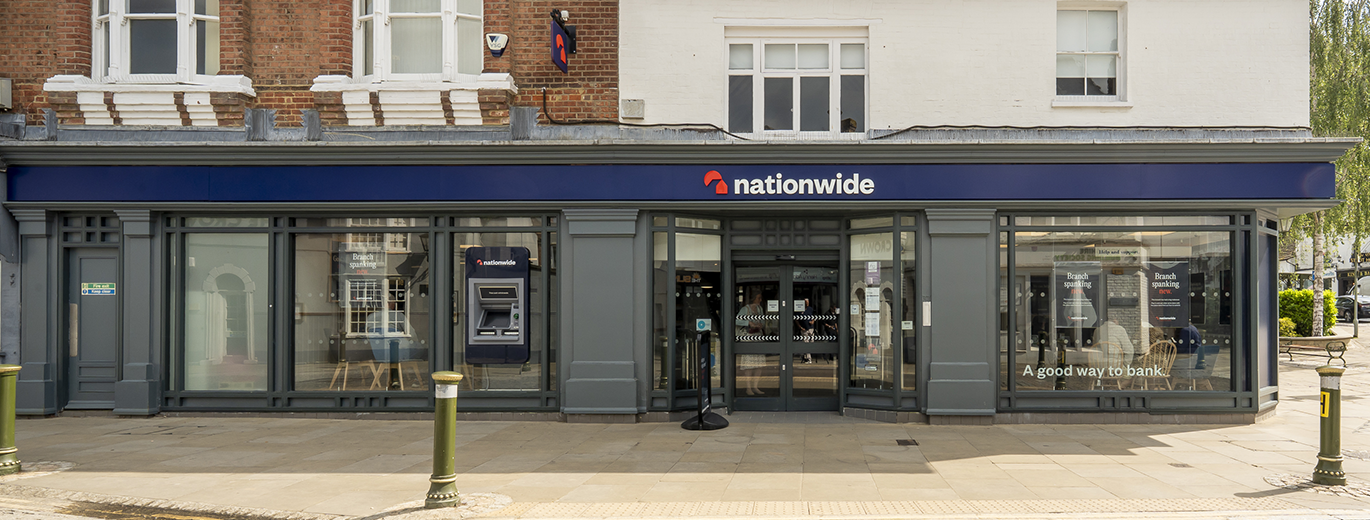 “Exterior view of a Nationwide bank branch with a blue sign displaying the Nationwide logo and name. The building has large glass windows, an ATM on the left si