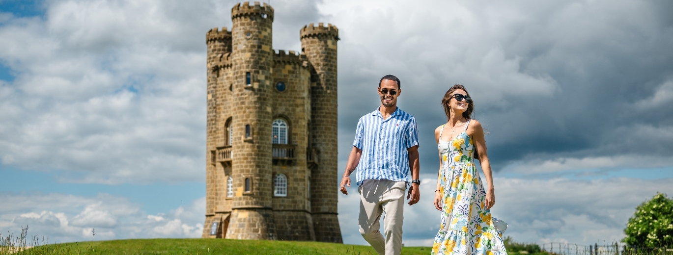 a white man and woman, stand in front of a sandstone turret, with brooding skies,  