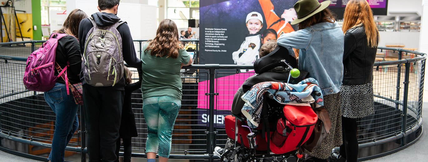 "A group of people looking across a barrier down to the next level with a tall poster in the middle, the writing on the poster is too small to read in the photo."