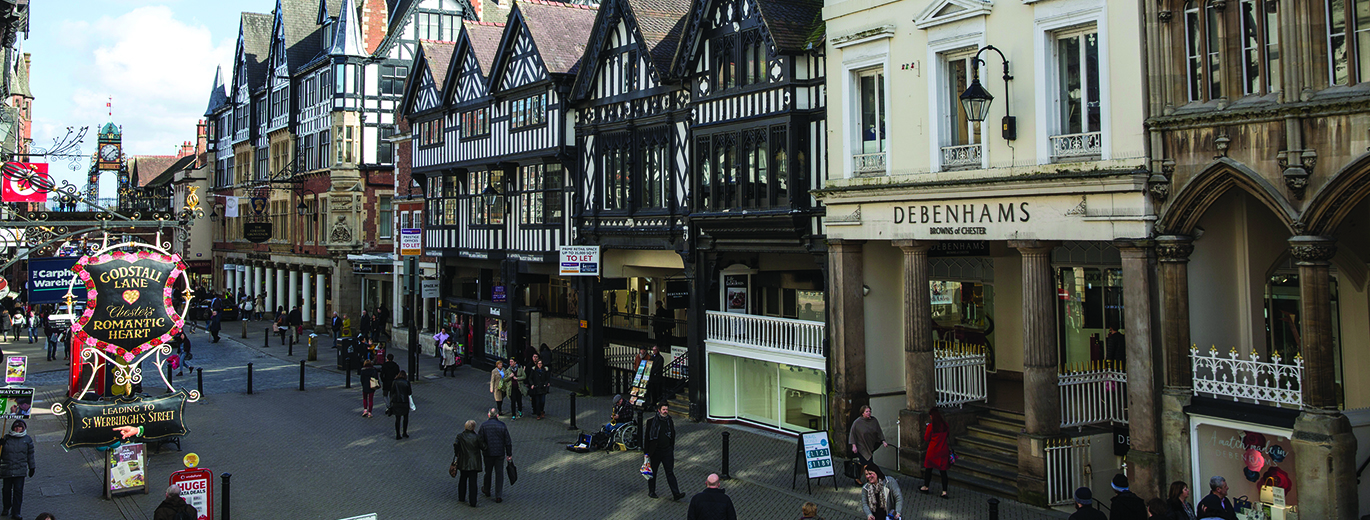 Busy pedestrianised street lined with historic black-and-white timber-framed buildings, including a “DEBENHAMS” storefront, with people walking, cyclists passing, and shop signs along the road.