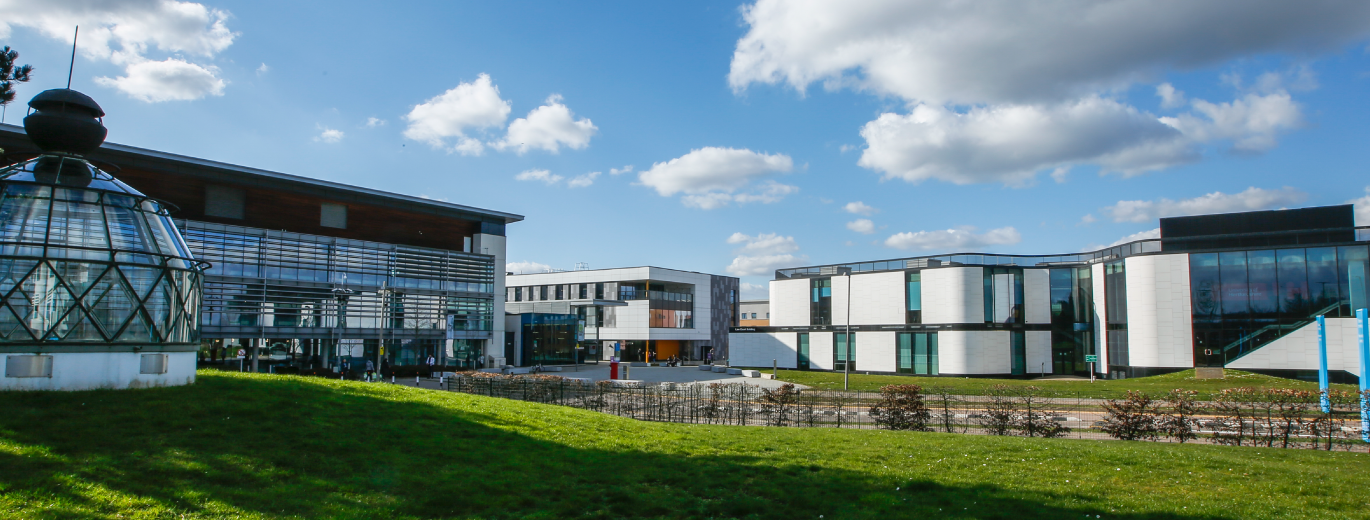"Academic buildings on College Lane Campus. The buildings are large and modern with glass and white panelling. In the foreground is a large patch of grass. Blue skies in the background."