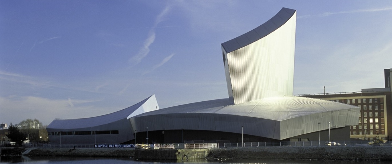 "Photo from across the river of the outside of IWM North. The building has a grey roof with angled features. There is a sign saying Imperial War Museum North to the left."