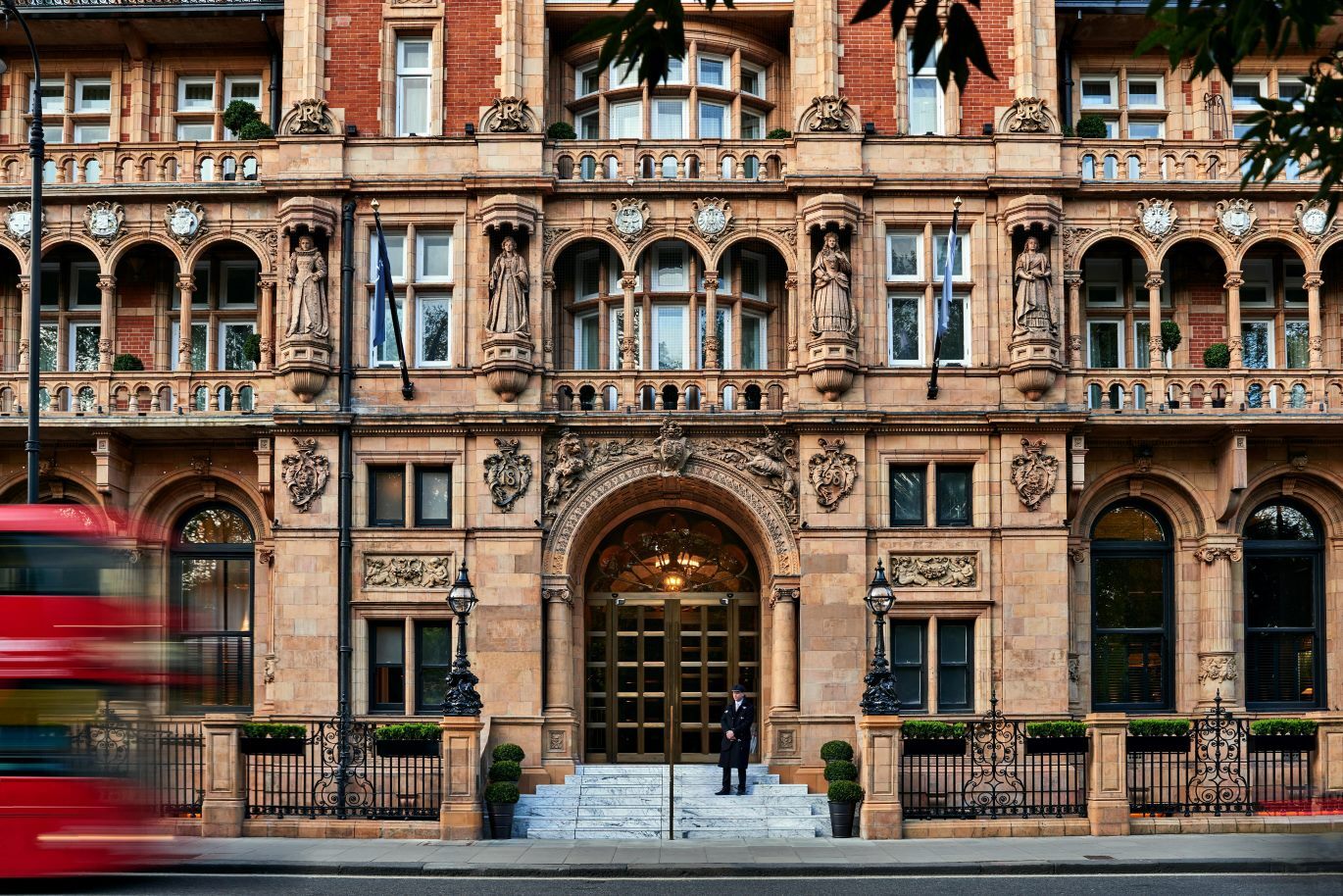 "Large brown sandstone hotel with arched doorway, railings and several stories can be seen with a red double decker bus at the edge of the image"