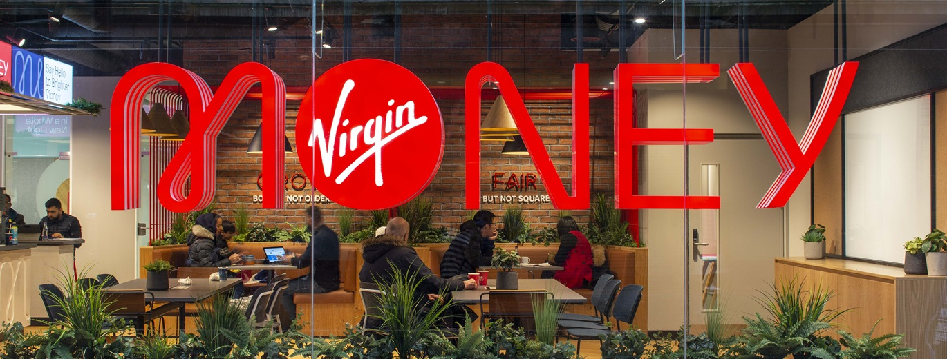 "Interior view of a modern Virgin Money branch with people sitting at tables, working and chatting. Large red “Virgin Money” branding is displayed across the glass window in the foreground, with plants and warm lighting creating a relaxed café-style atmosphere"
