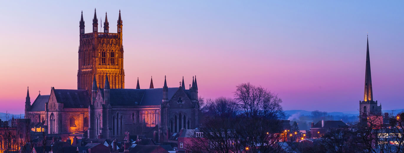 Worcester Cathedral illuminated at dusk, with its tower and spires rising above the city skyline against a pink and purple evening sky.