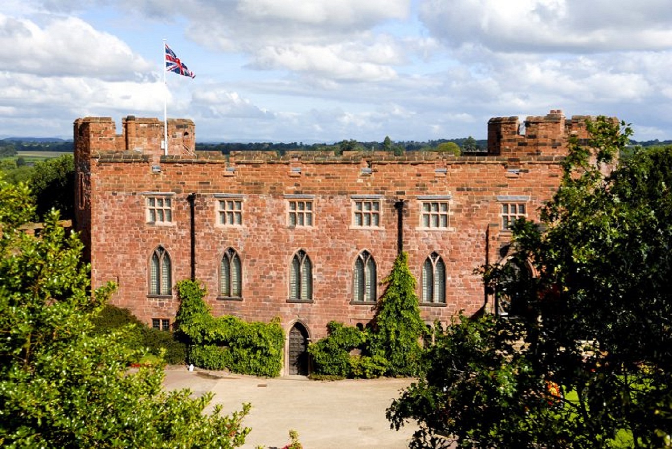 Red sandstone castle with crenellated towers and narrow arched windows, set on a hill with trees in the foreground and a flag flying on top.