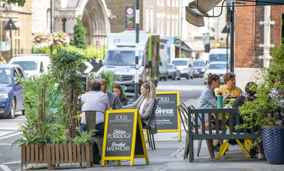 Outdoor café seating on a busy city street, with people sitting at tables beside potted plants and passing traffic, and a yellow chalkboard sign reading “COFFEE & CAKE.”