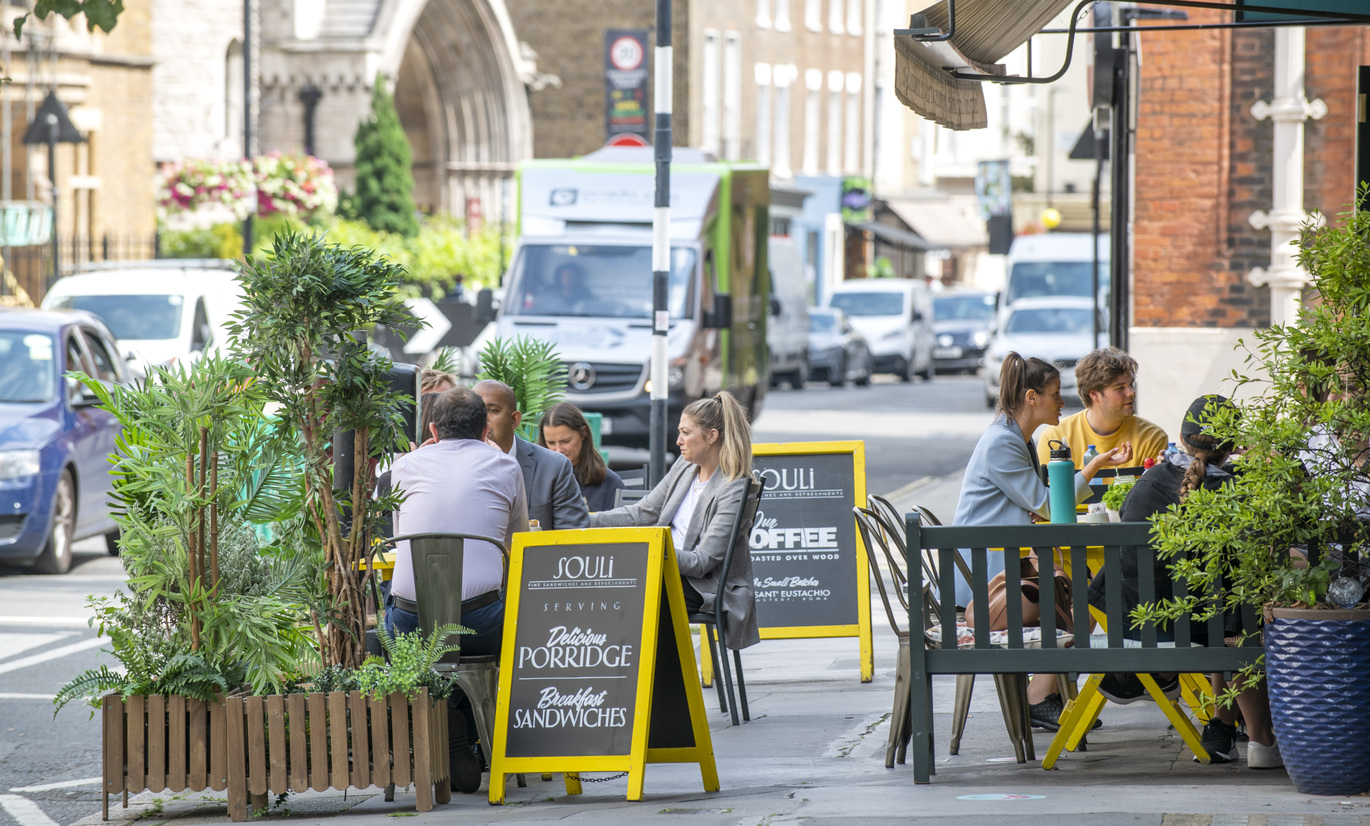 Outdoor café seating on a busy city street, with people sitting at tables beside potted plants and passing traffic, and a yellow chalkboard sign reading “COFFEE & CAKE.”