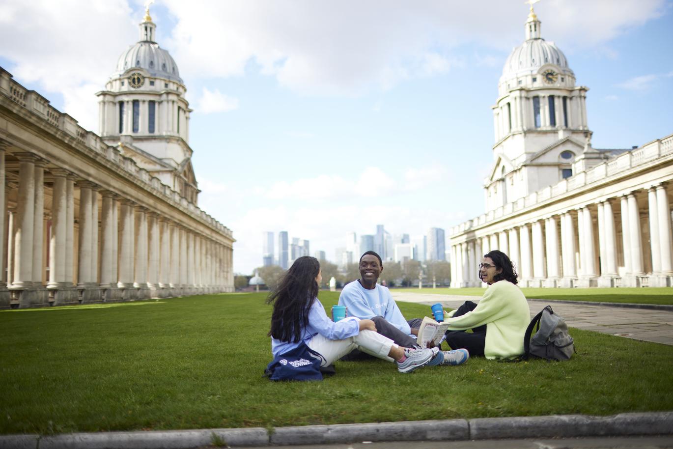 "Three students sit on a grassy lawn between two historic colonnaded buildings with domed towers, facing each other in a small group. A modern city skyline is visible in the distance under a bright, partly cloudy sky"