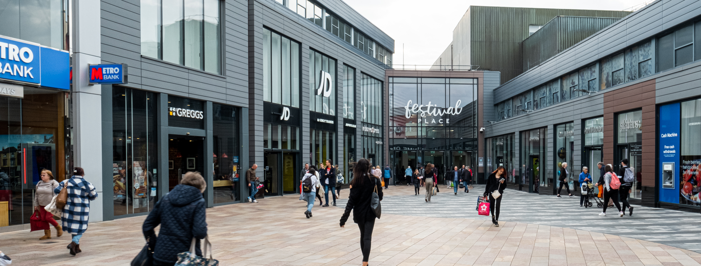 "Festival Place Shopping Centre entrance showing Metro Bank, Greggs, JD Sports and Vision Express on the left and a cash machine, Slaters, Card Factory and another store entrance on the right"                                                                                             