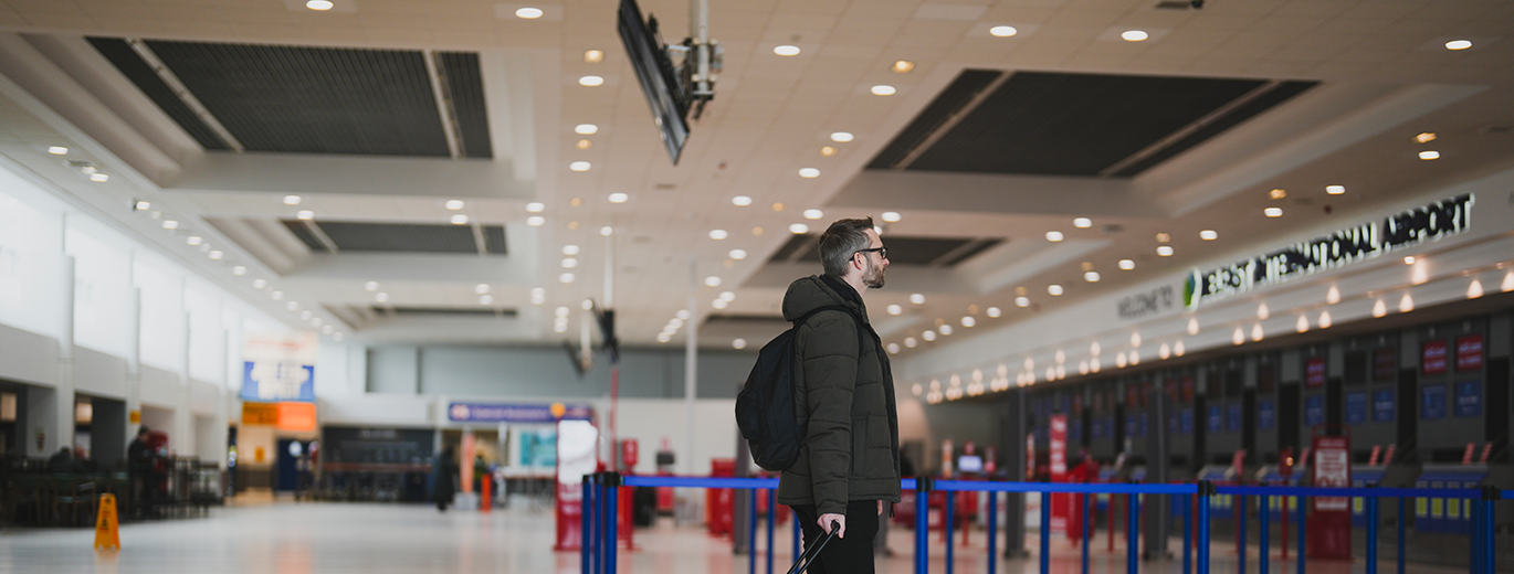 "Photograph shows a white man with dark hair and beard, wearing glasses and black clothing with luggage, looking towards the Belfast International Airport check in desks"